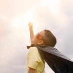 A young boy dressed in a cape and sunglasses, with arm outstretched to the sky.