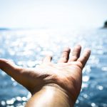 A picture of a hand, palm upward, outstretched towards the horizon, with an ocean in the background..
