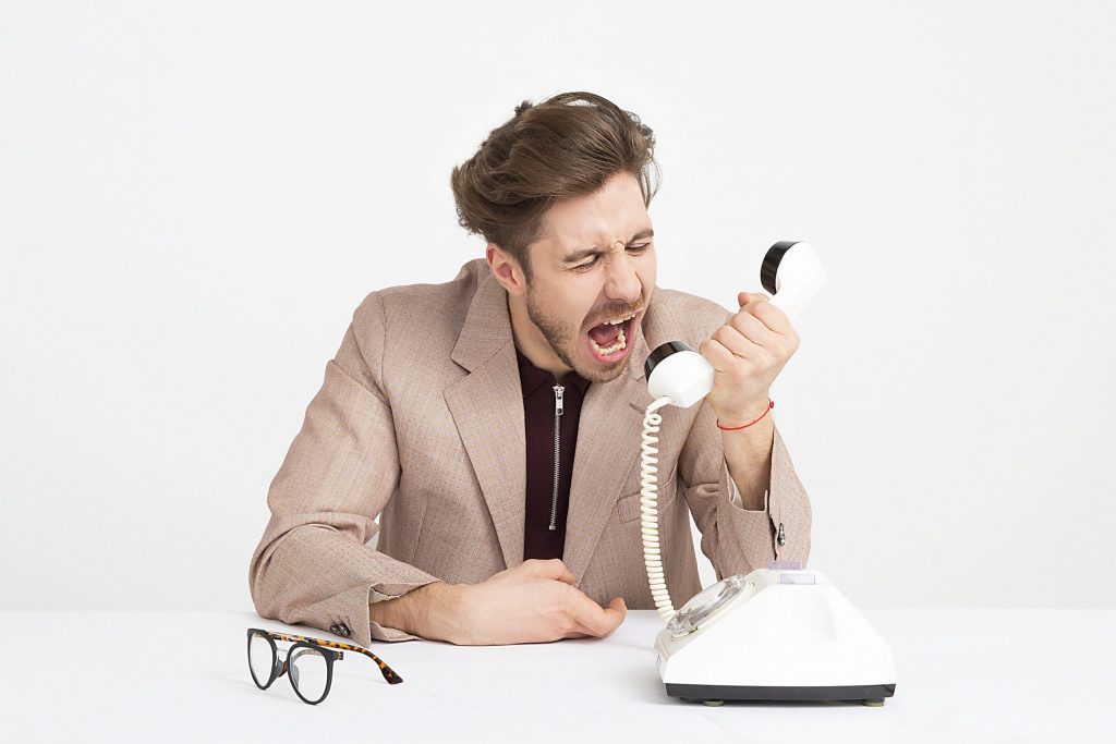 A man yelling into an old-style white telephone, with glasses sitting on the table in front of him