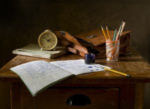 A wooden desk. Sitting on top are a copy book with a fountain pen and ink, a glass jar holding other pens and pencils, a notebook, clock and satchel.