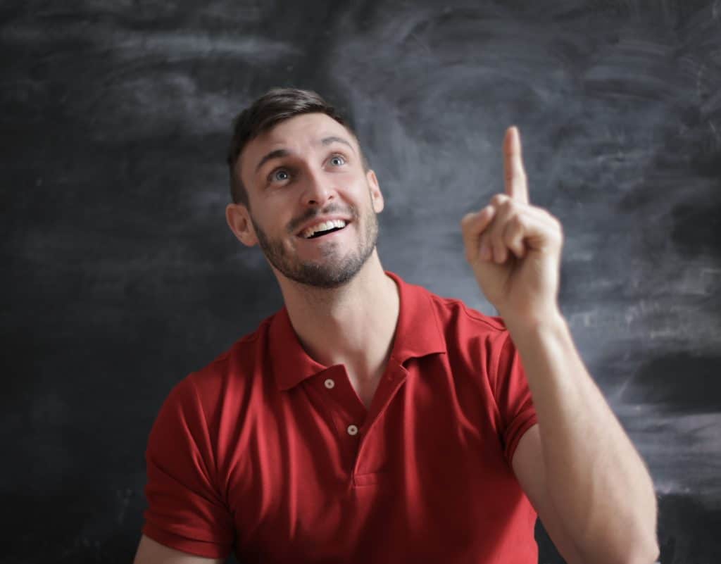 A man in a red shirt in front of a black background. He is holding his left arm in the air with the index finger raised, looking upward and smiling as though he has had an insight.
Image credit: Pexels