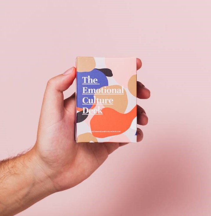 A hand holding a pack of The Emotional Culture Deck cards against a pink background