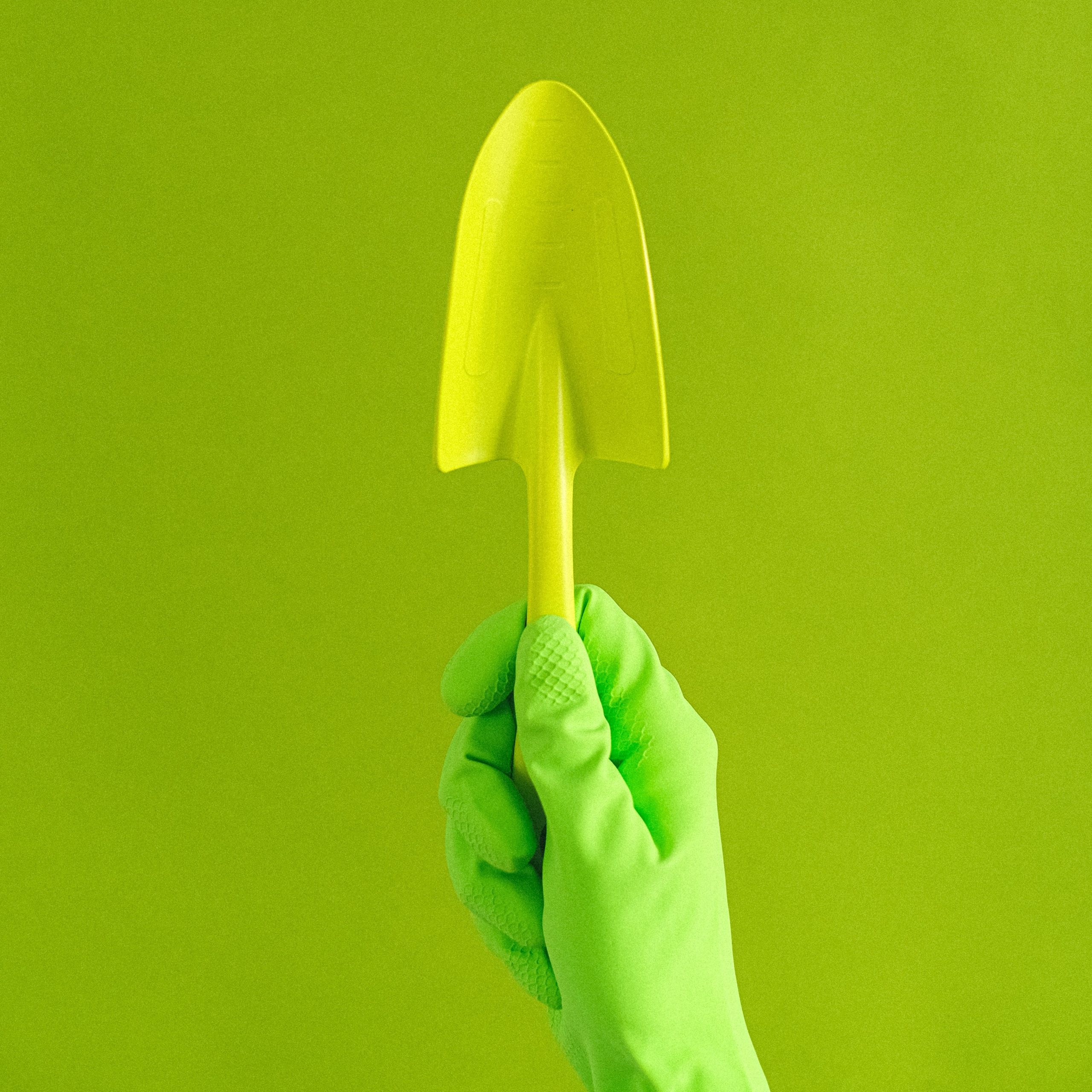 A photo of a green-gloved hand holding a green garden trowel against a green background