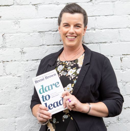 Photo of Daria Williamson leaning against a white wall, holding the book 'Dare to Lead' by Brené Brown