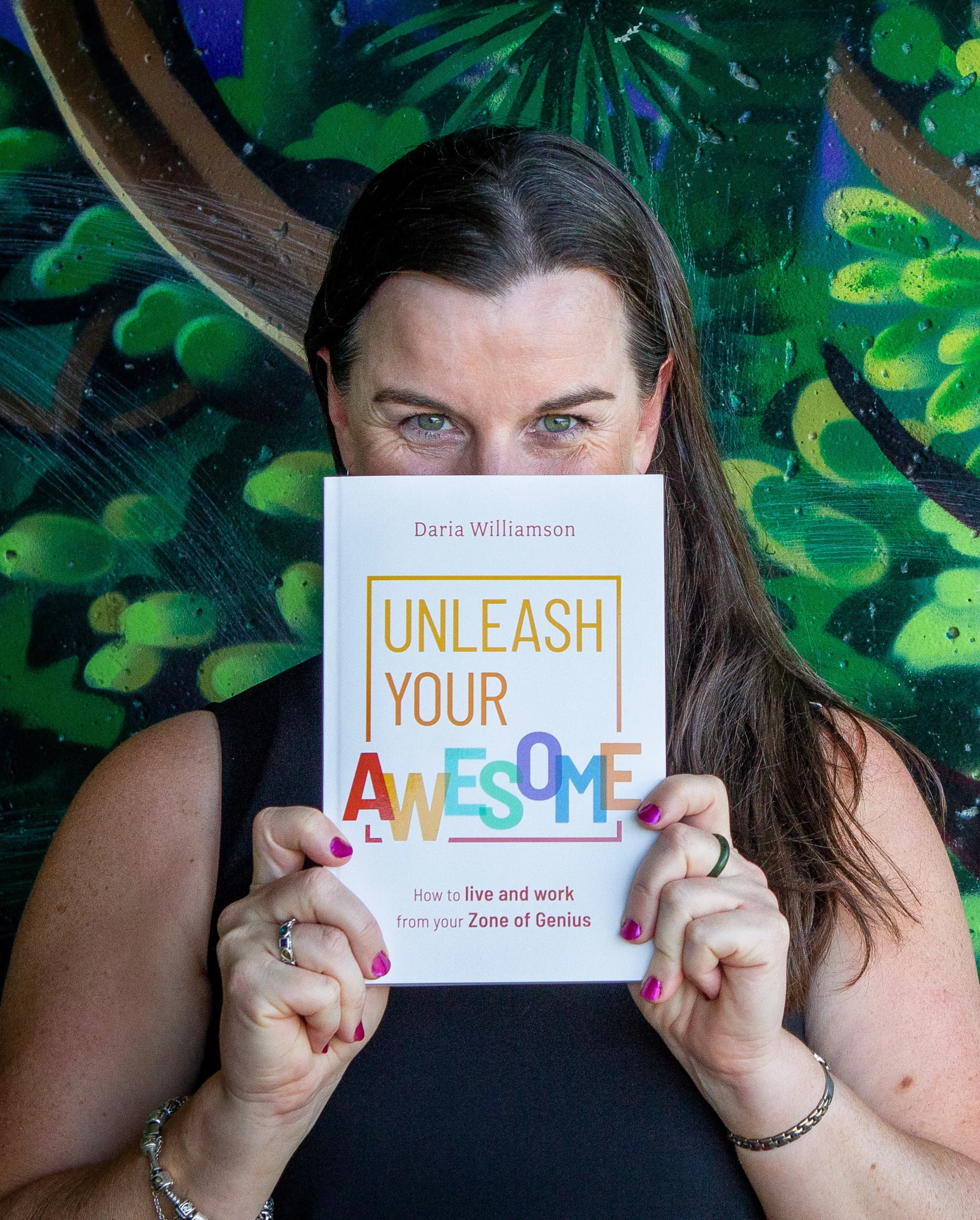 A dark-haired woman stands in front of a concrete wall that has been painted to resemble a jungle. She is holding a copy of the book 'Unleash Your Awesome' in front of her face, so that only her eyes are peeking out - she appears to be smiling broadly.
