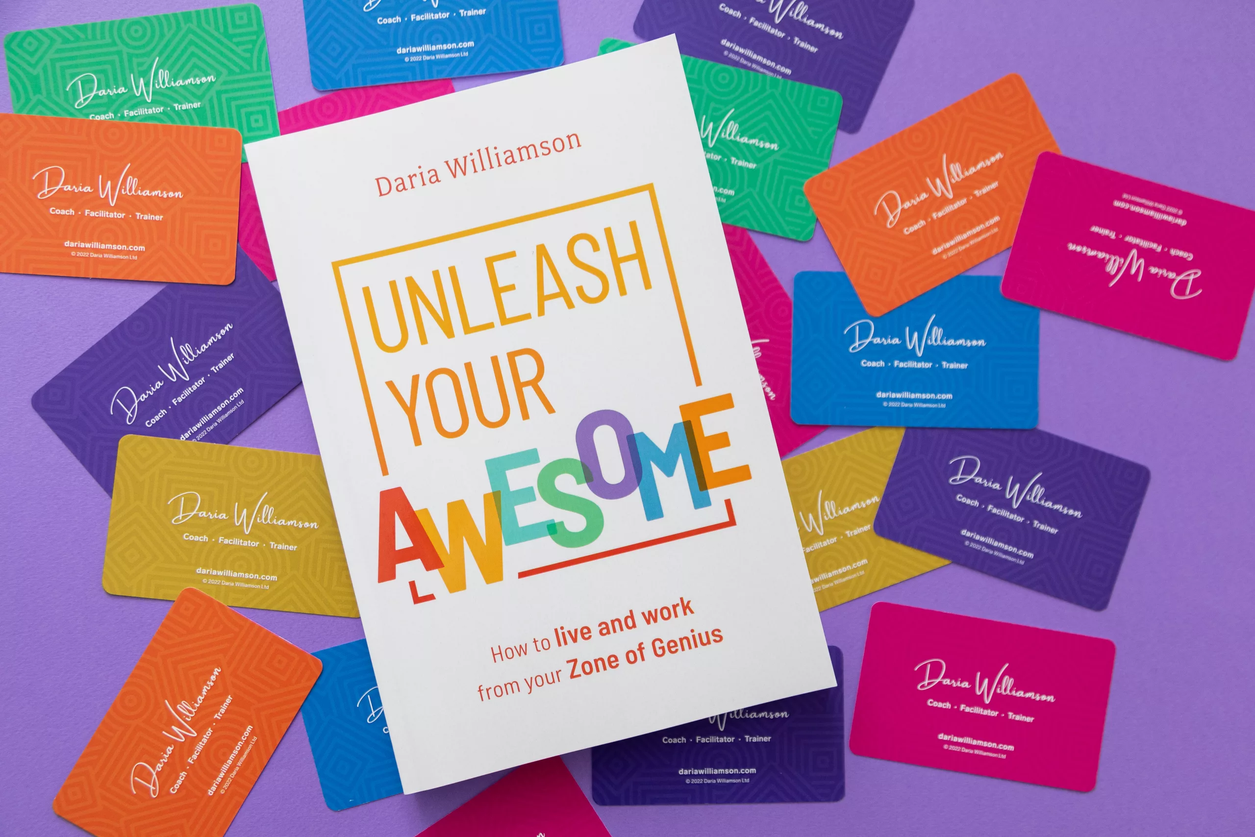 A copy of the book 'Unleash Your Awesome' lies atop a sample of The Strengths Deck cards, which have been scattered face-down on top of a purple background, so that the colourful backs of the cards are visible.