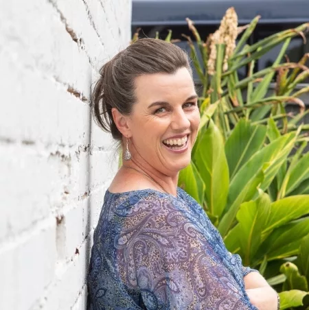 A dark-haired woman wearing a blue paisley top is leaning against a brick wall painted white. Her head is turned to look over her right shoulder, and she has a broad, open-mouthed smile, as though she has been caught laughing. In the background is a garden and dark wall