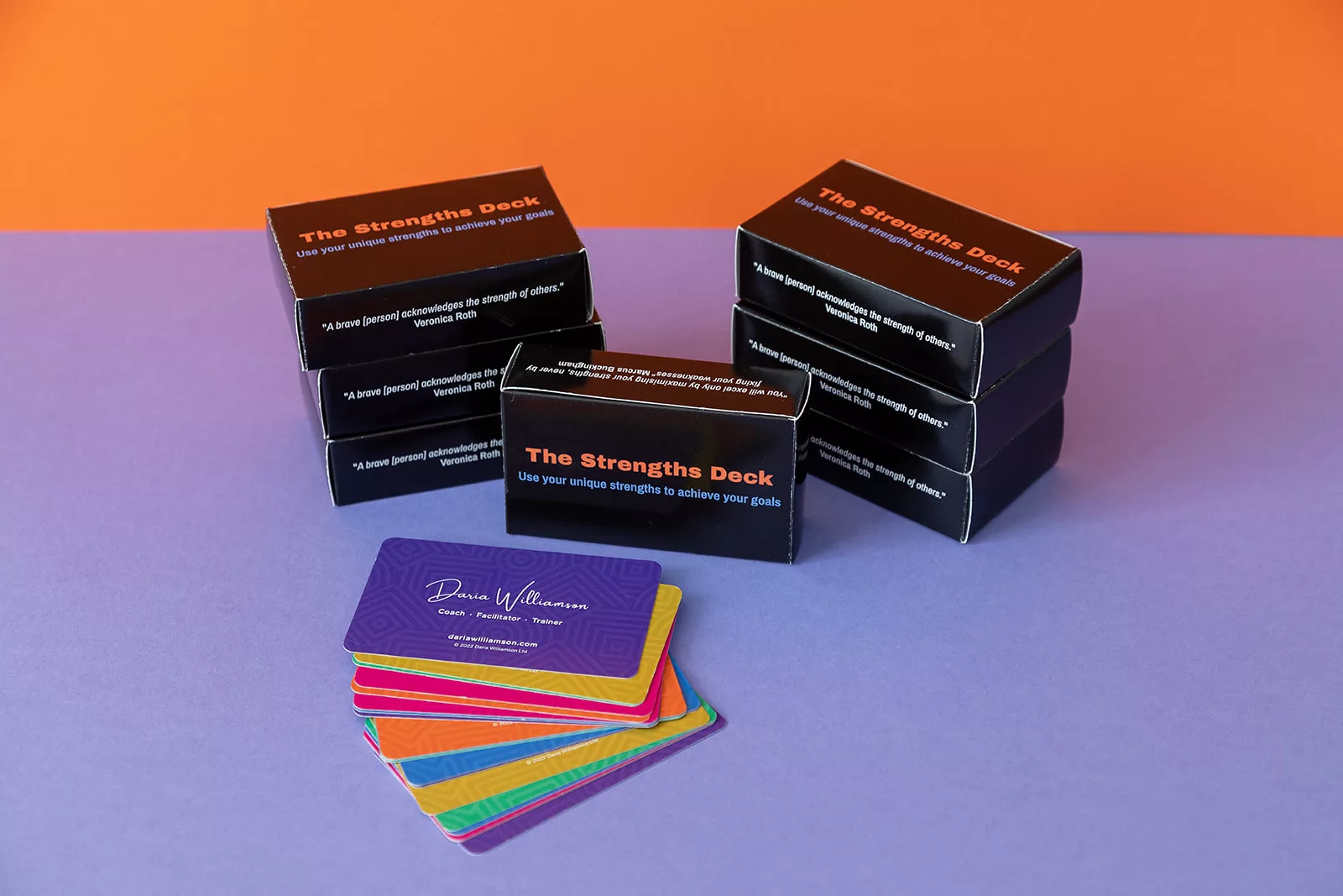A stack of black boxes with "The Strengths Deck" logo on them are stacked on a purple surface, with an orange background. In front of the stacks of card boxes, a handful of brightly-coloured cards are fanned out, showing the Daria Williamson logo.