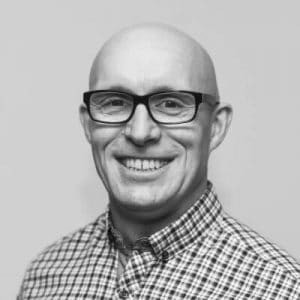 Black and white photo. Karl Craig-West is standing in front of a pale background. He is bald, wears glasses and a checked shirt, and is smiling broadly at the camera.