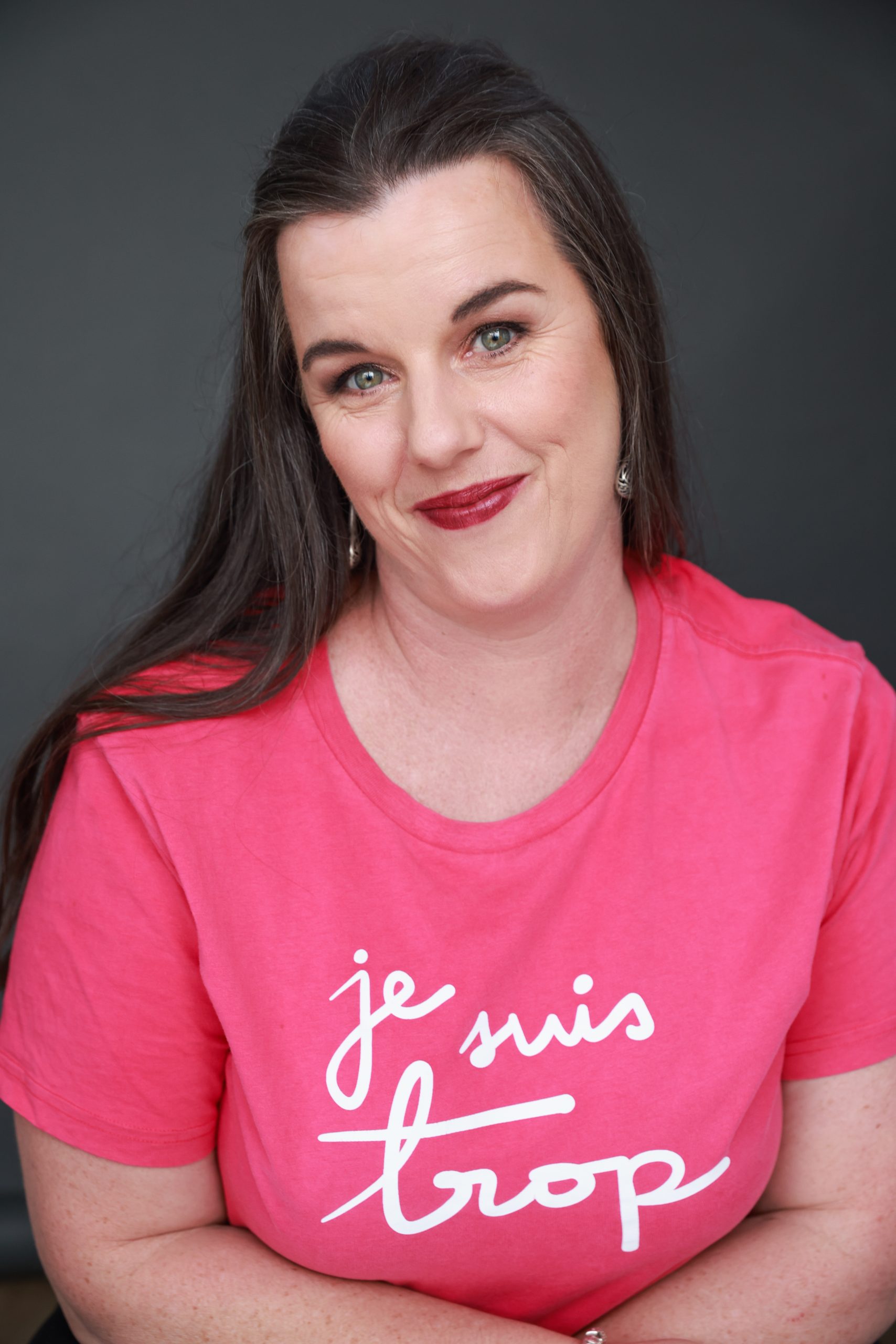 Daria, a dark-haired woman is wearing dark red lipstick and a bright pink top with the words 'je suis trop' (I am too much) in white lettering. She is looking directly at the camera, with her head tilted, and a slightly exasperated expression.
