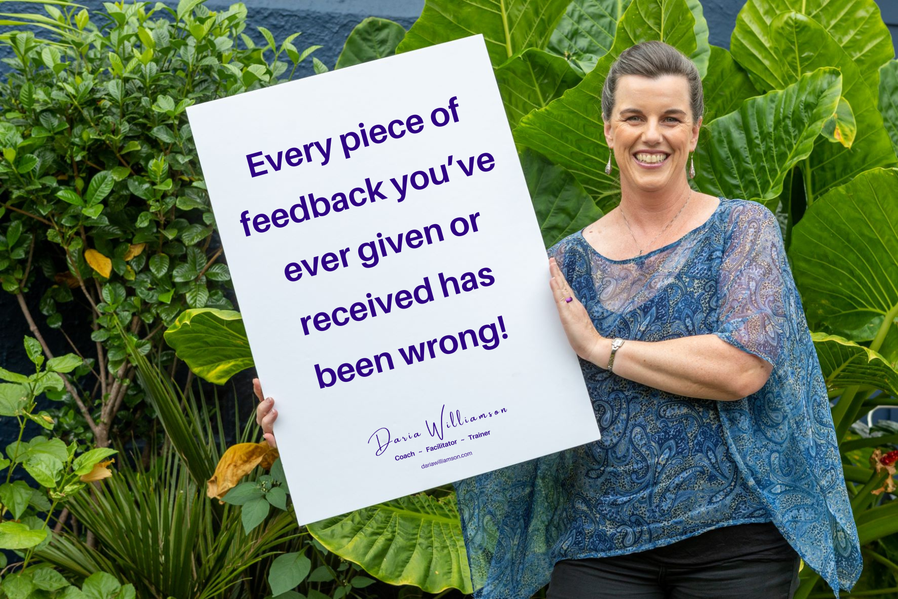 A dark haired woman standing in front of a leafy background with a sign that has the text "every piece of feedback you've ever given or received has been wrong!"