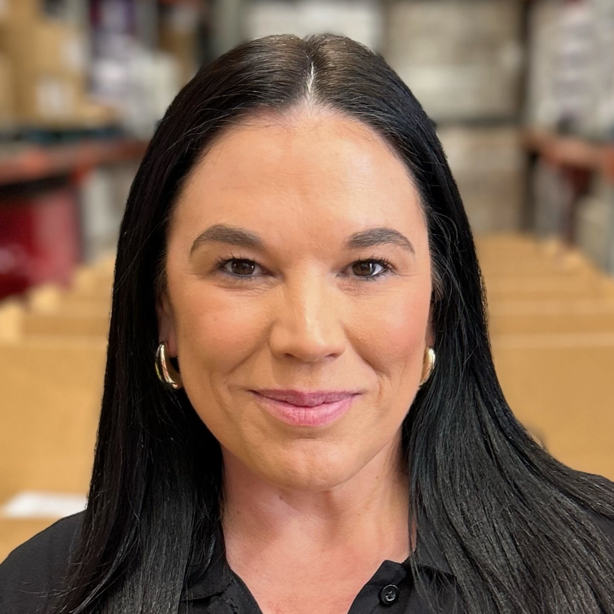 Sarah Page, a dark-haired woman is standing in a warehouse. She wears a black long-sleeved shirt, and is smiling at the camera.