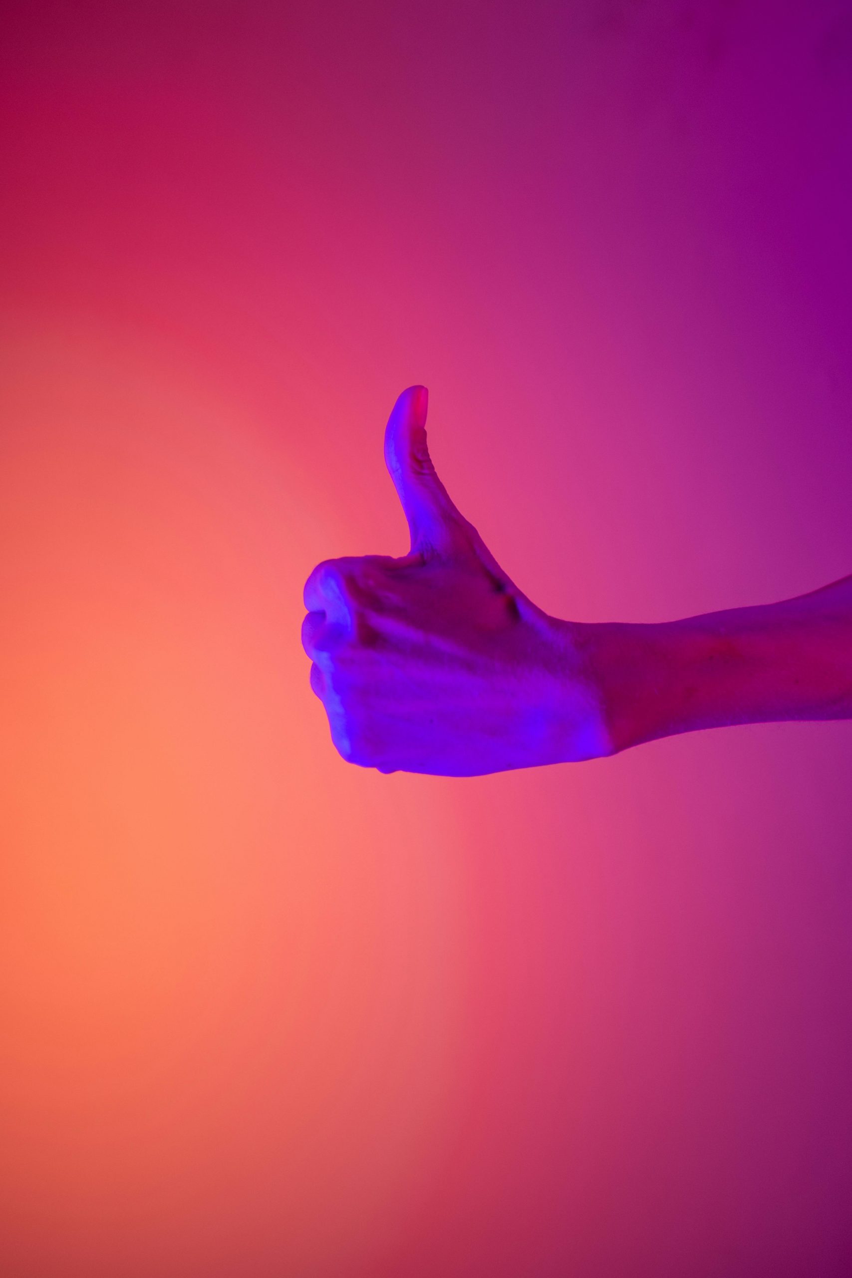 An image of one hand lit with purple light against an orange background. The hand is giving a "thumbs up" sign.