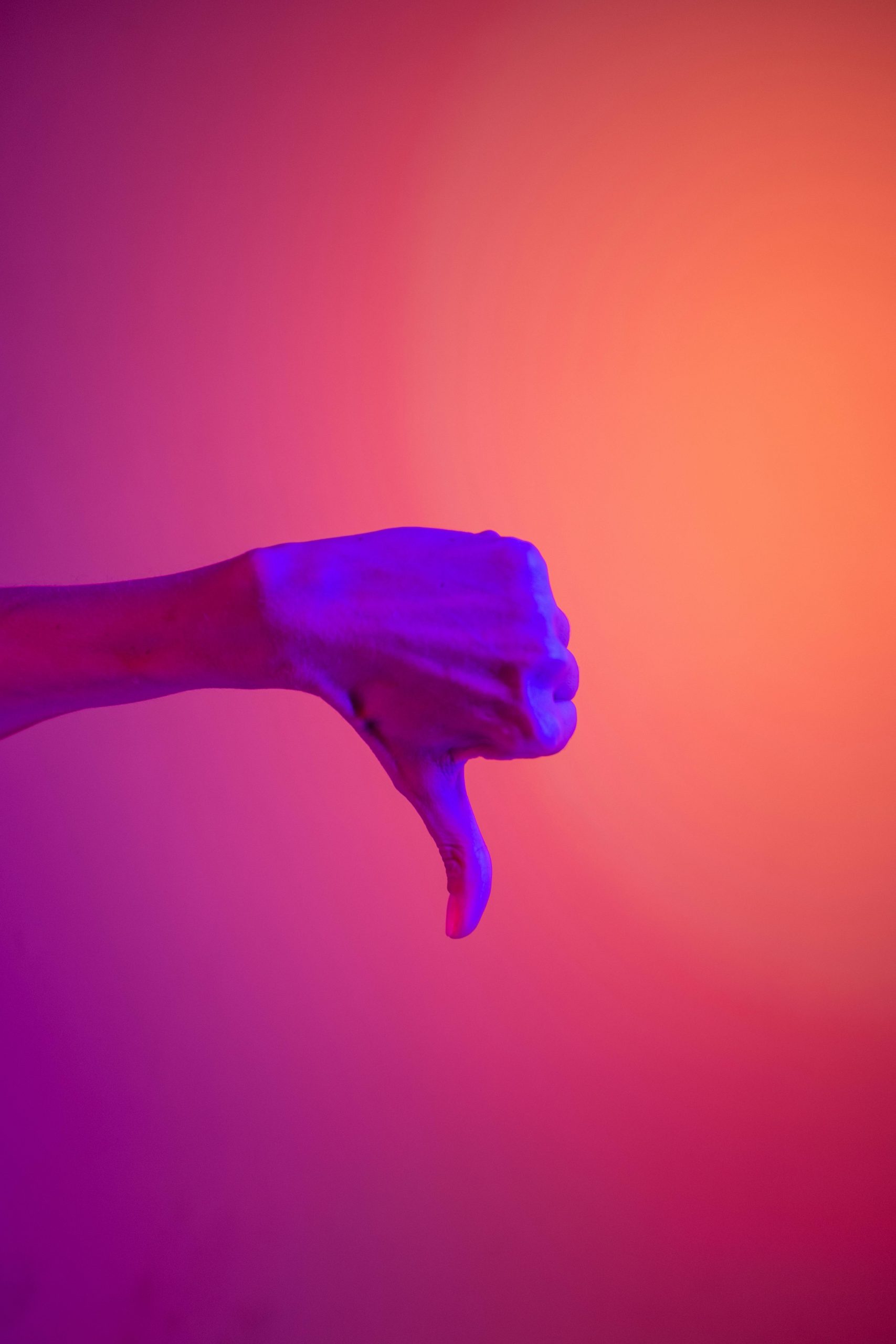 An image of one hand lit with purple light against an orange background. The hand is giving a "thumbs down" sign.