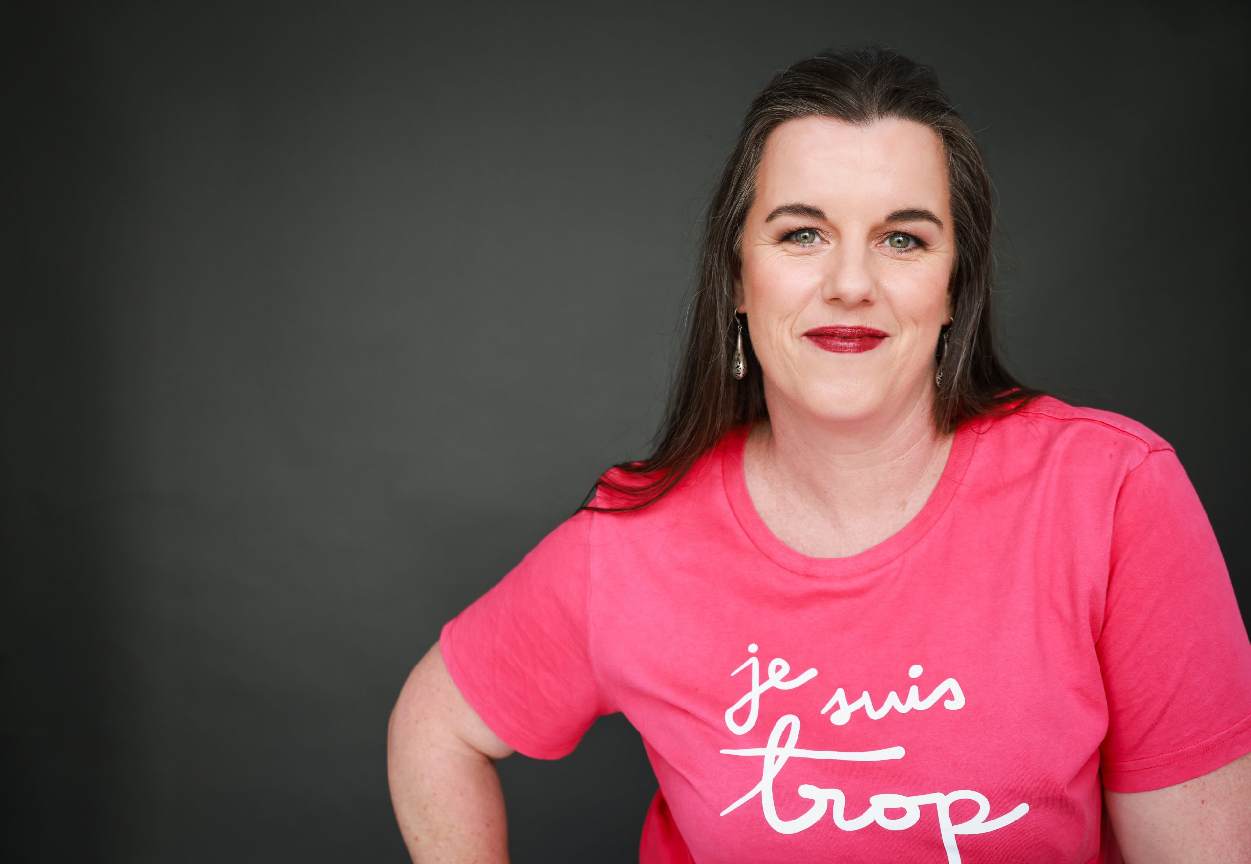 Daria, a dark-haired woman is wearing dark red lipstick and a bright pink top with the words 'je suis trop' (I am too much) in white lettering. She is looking directly at the camera, with a hand on one hip, smiling.