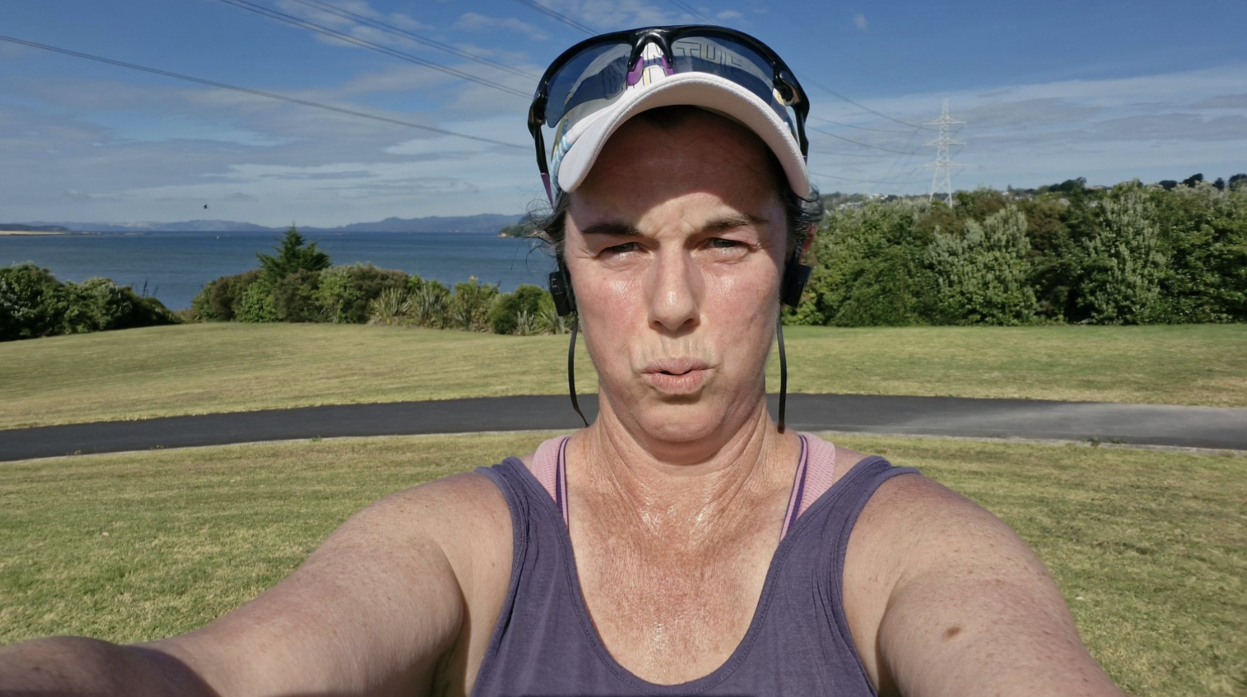 A dark-haired woman in workout clothing, with a baseball cap and sunglasses on top of her head, is squinting at the camera and exhaling hard. Behind her, in the near background, is grass, a pathway, and a line of trees. In the middle and far distance is a harbour and hills.