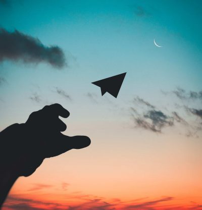 Silhouette photo of hand throwing a paper plane into a dusky sky, towards a crescent moon