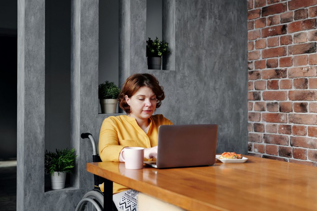 A woman in a wheelchair, working at a dining table with a laptop, coffee mug and snack.