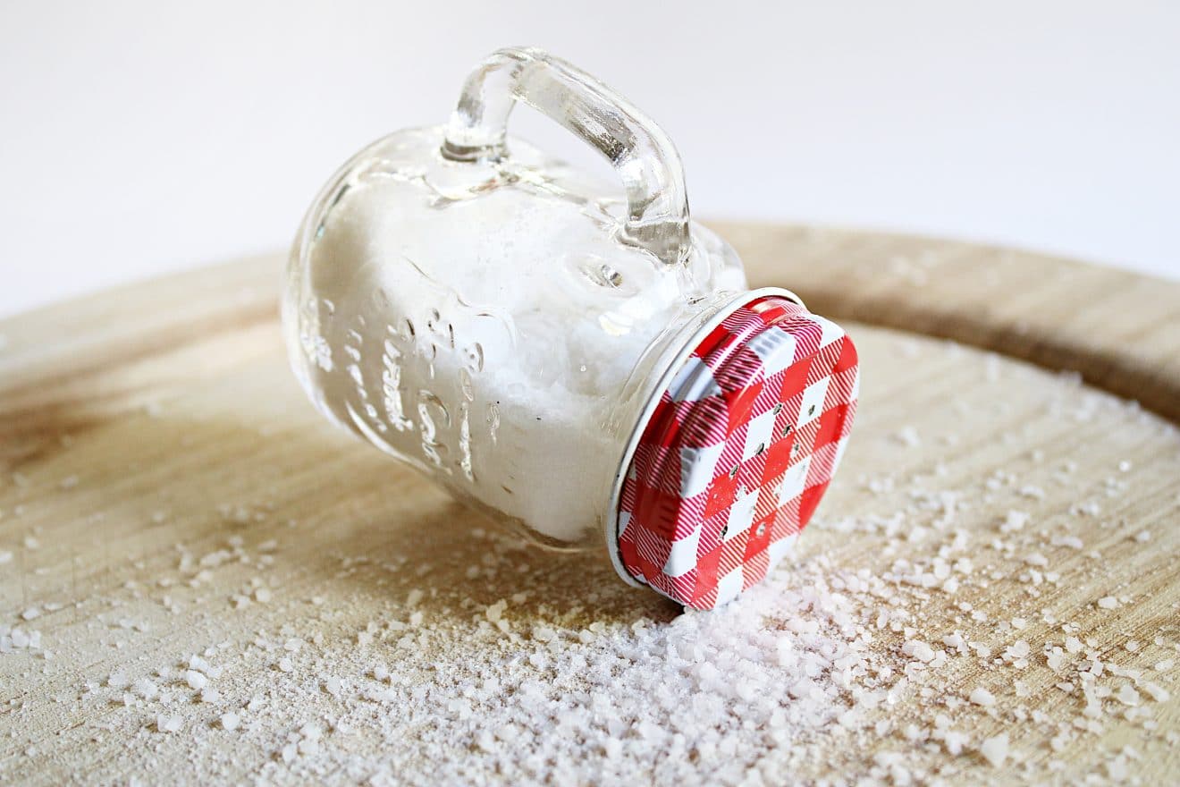 A glass container of salt with a red lid, lying on its side on a wooden board, with salt crystals sprinkled around