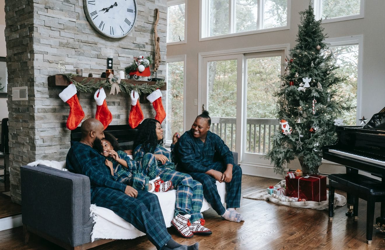 A family sitting on a couch in front of a fireplace with stockings on the mantle, and a Christmas tree by the window