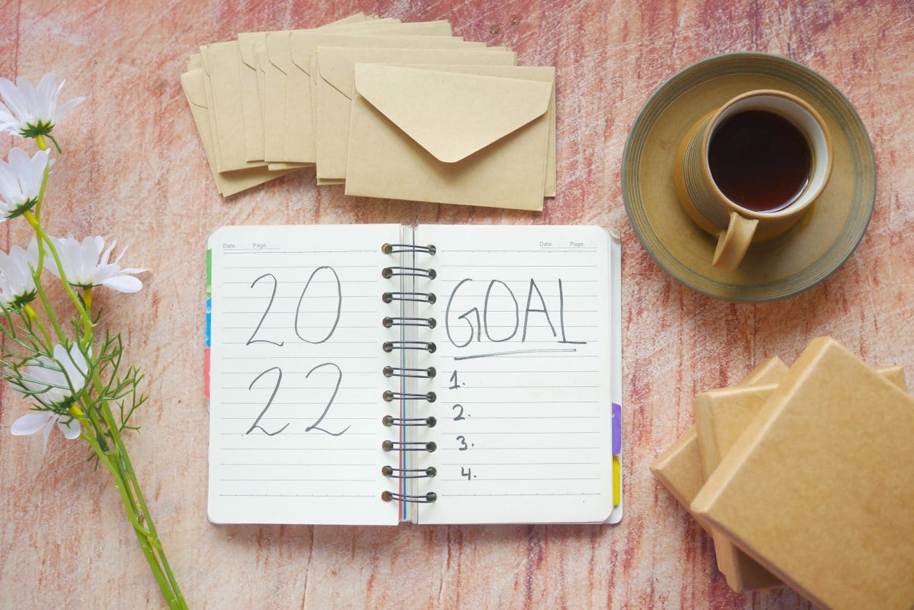 An overhead photo of a desk, with flowers on the left, a diary in the middle with "2022" on the left page, "Goal, 1, 2, 3, 4" on the right page, a set of envelopes above the diary, and a coffee cup and boxes to the right