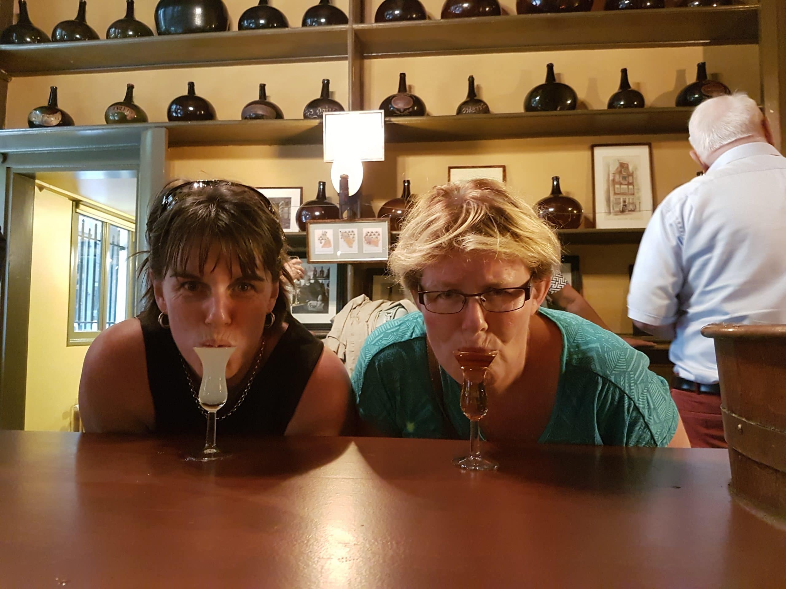 Photo of two women, one dark-haired, one fair-haired, in a bar, learning forward to sip from the liqueur glasses in front of them