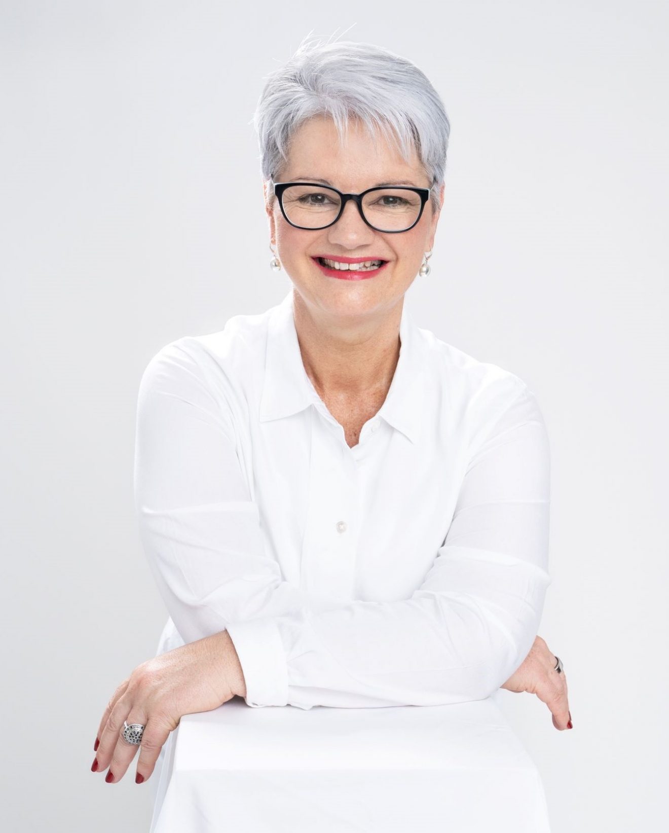 Photo of Deb Bailey, a smiling, silver-haired woman with glasses, wearing a white shirt and leaning forward on a white box in a relaxed posed