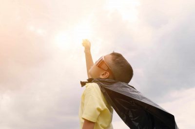 A young boy dressed in a cape and sunglasses, with arm outstretched to the sky.
